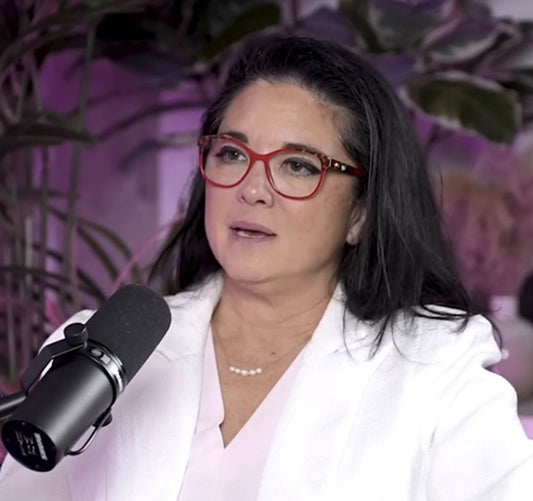 Dr. Vonda Wright speaking into a podcast microphone wearing red glasses and a white blazer, discussing women’s bone health.
