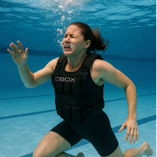 A woman wearing a black weight vest struggles underwater in a swimming pool, with bubbles rising around her and an expression of effort on her face, illustrating the danger and absurdity of trying to swim with a weight vest